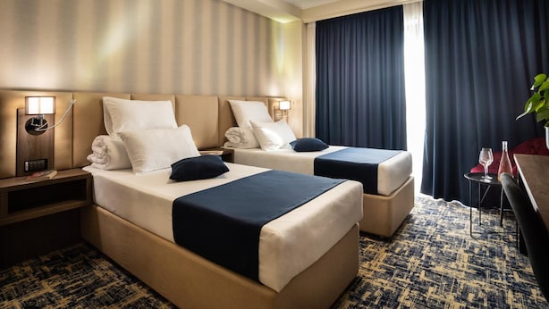 A hotel room with two beds, a nightstand with a lamp, a small table with a glass and a bottle, and a large window with curtains.