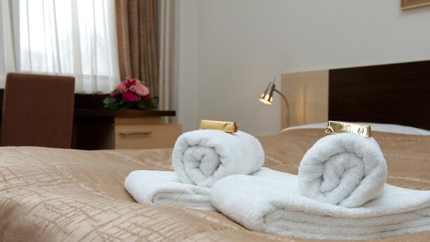Una cama bien tendida con dos toallas blancas enrolladas y un sujetador dorado, una mesita de noche de madera con una lámpara, y una ventana con cortinas.
