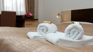 A neatly made bed with two rolled white towels and a gold clip, a wooden nightstand with a lamp, and a window with curtains.