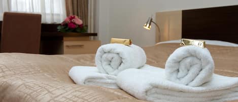 Cama bem arrumada com duas toalhas brancas enroladas e um prendedor dourado, mesa de cabeceira de madeira com luminária e janela com cortinas.