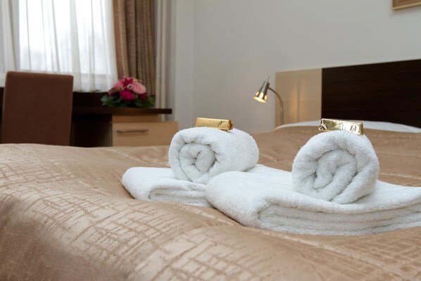 Cama bem arrumada com duas toalhas brancas enroladas e um prendedor dourado, mesa de cabeceira de madeira com luminária e janela com cortinas.