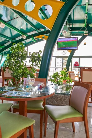 A modern indoor space with a glass ceiling, green plants, a round table with chairs, and a television screen. - Green Center Gili (Gostivar)