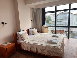 A bedroom with a large bed, a bedside table, a wall-mounted lamp, and a window with a view of buildings.