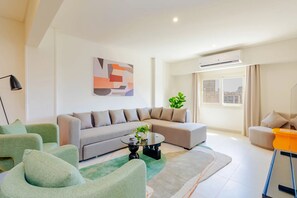 A modern living room with a grey sectional sofa, a green armchair, a round coffee table, and a large window with a view of the city. - Brassbell L Zamalek L Chroma L Central Location (Cairo)