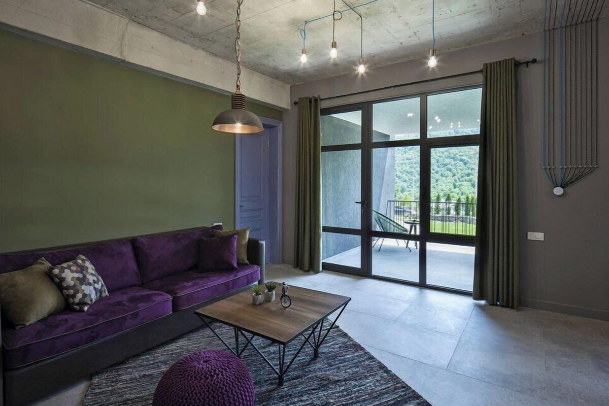 A modern living room with a purple sofa, a wooden coffee table, and a large sliding glass door leading to an outdoor area.
