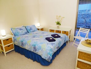 Un dormitorio con cama, mesitas de noche, una silla, una pequeña mesa y una ventana con vistas.