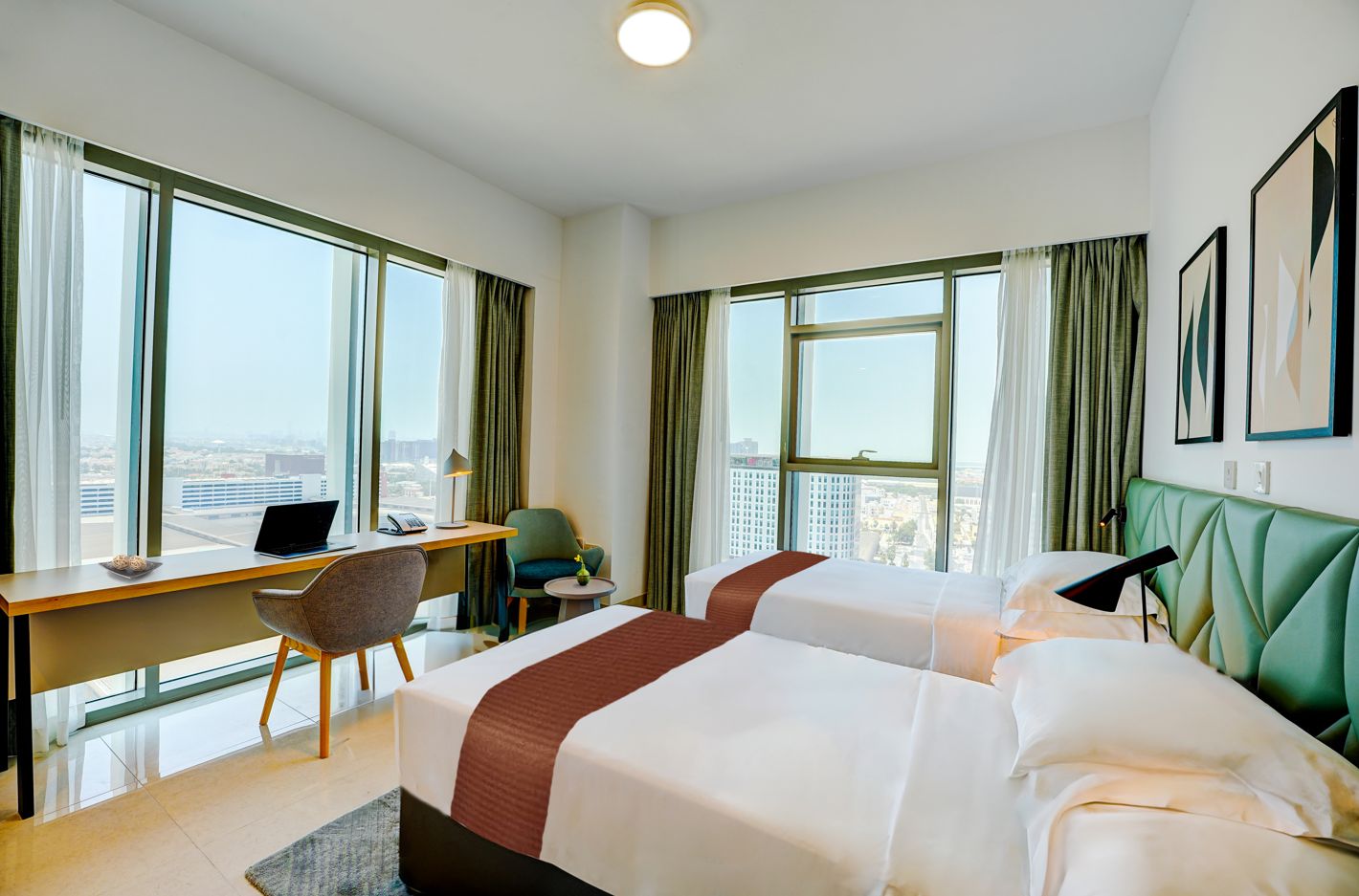 Ett hotellrum med två sängar, ett skrivbord, en stol och stora fönster med gardiner.