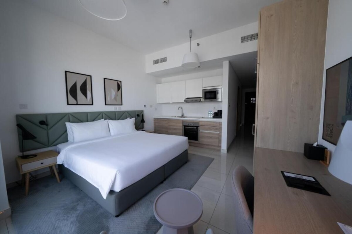 A modern hotel room with a large bed, a desk, a chair, a small table, a microwave, and a sink in the adjacent kitchenette.
