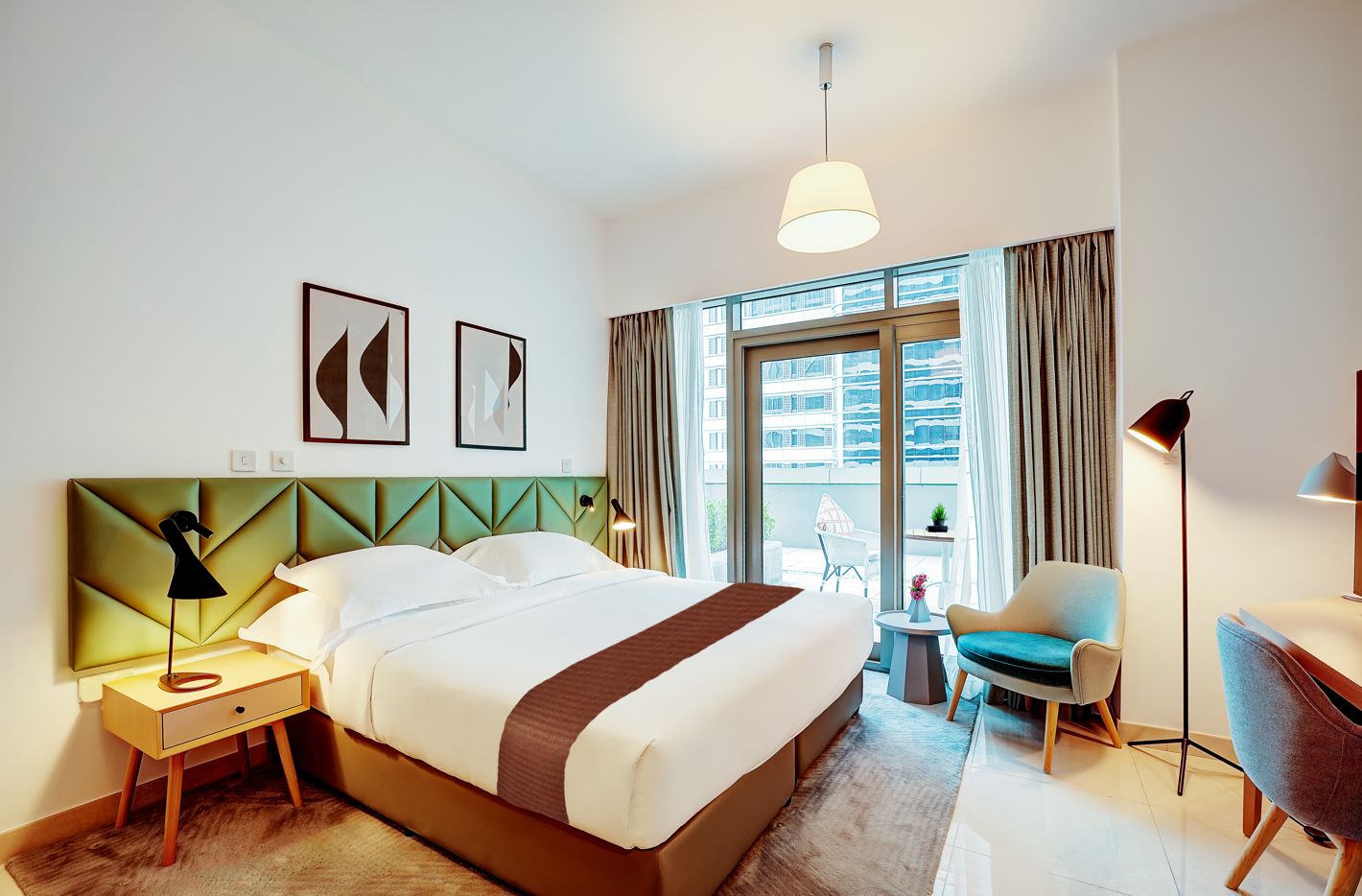 Ett hotellrum med en säng, sängbord, ett skrivbord, en stol, en lampa, ett fönster med gardiner och konstverk på väggen.