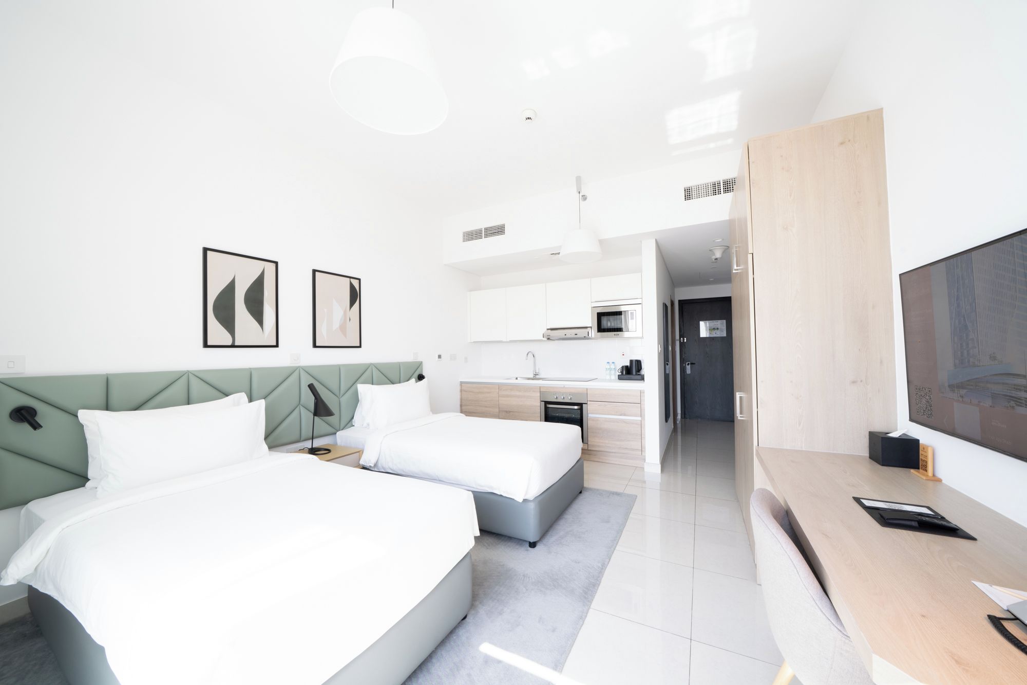A modern hotel room with a bed, bedside tables, a desk, a TV, and a kitchenette.