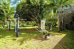 Equipos de ejercicio al aire libre, un edificio de piedra y exuberante vegetación en un parque.
