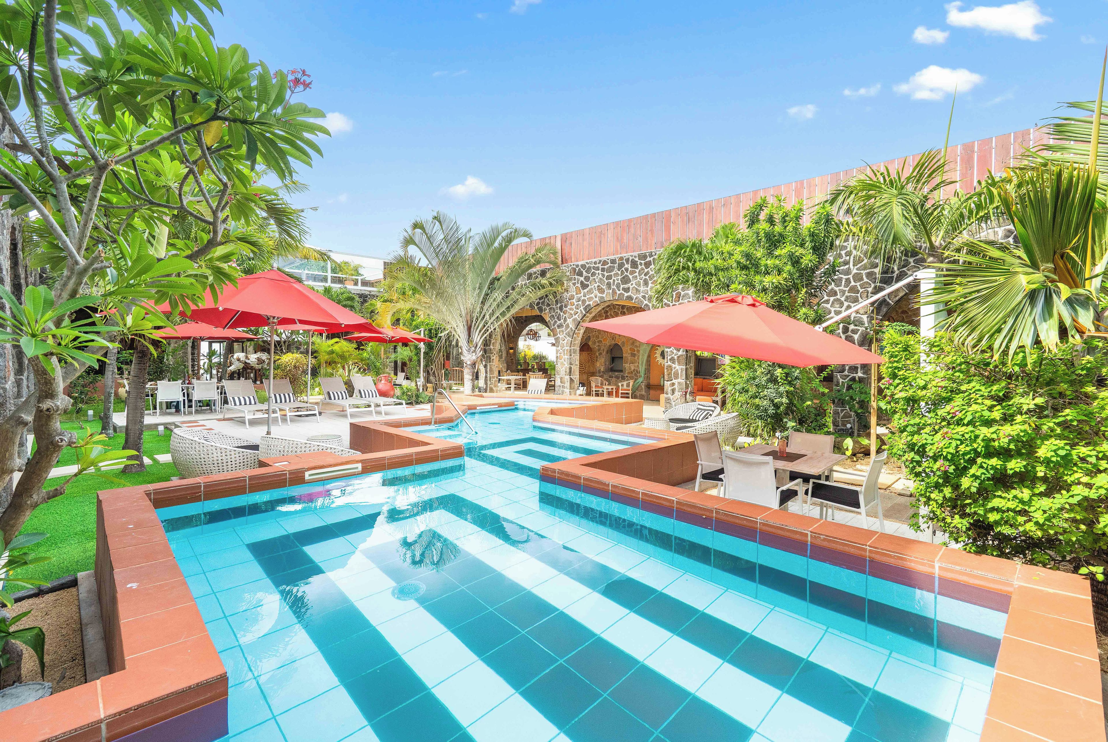 Piscina con bordes de azulejos, rodeada de sillas de descanso y sombrillas rojas, con un fondo de vegetación y arquitectura de piedra.