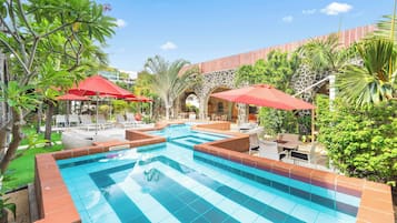 Piscina con bordes de azulejos, rodeada de sillas de descanso y sombrillas rojas, con un fondo de vegetación y arquitectura de piedra.