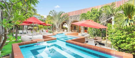 Piscina con bordes de azulejos, rodeada de sillas de descanso y sombrillas rojas, con un fondo de vegetación y arquitectura de piedra.