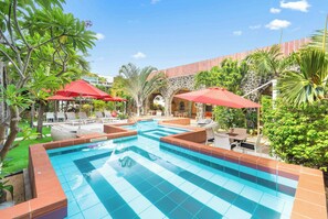 Piscina con bordes de azulejos, rodeada de sillas de descanso y sombrillas rojas, con un fondo de vegetación y arquitectura de piedra.