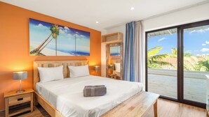 Habitación de hotel con cama, mesitas de noche, un banco, un cuadro de una palmera y vista a palmeras a través de una puerta corrediza.