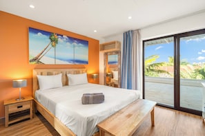 Habitación de hotel con cama, mesitas de noche, un banco, un cuadro de una palmera y vista a palmeras a través de una puerta corrediza.