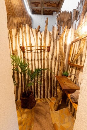 A rustic bathroom with a wooden slat wall, a hanging towel rack, a potted plant, a wooden stool, and a stone floor.