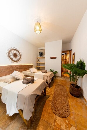 A hotel room with two beds, a woven rug, a fan, and a potted plant.