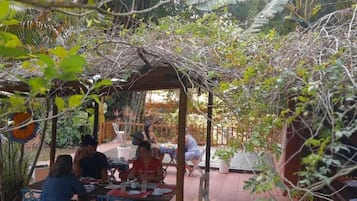 Uma área de pátio ao ar livre com mesas e cadeiras, cercada por vegetação e uma estrutura de pérgola.