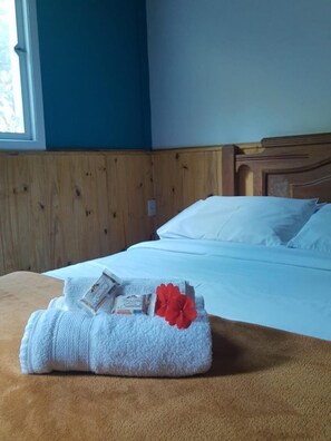 A neatly made bed with white linens and pillows, a brown blanket, and a folded towel with a red flower on a wooden headboard in a room with blue walls. - Pousada Casa Azul (Nova Friburgo)
