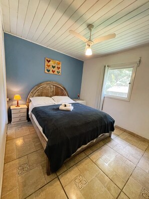 Un dormitorio con cama, mesita de noche, lámpara, ventilador de techo, ventana y un cuadro con diseño de corazones en la pared.