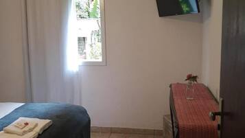 Quarto com cama, mesa com toalha vermelha, televisão na parede e janela com cortinas.