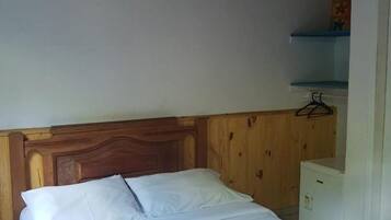 Uma cama de solteiro com cabeceira de madeira, um ventilador de teto e uma mesinha de cabeceira branca com uma toalha e um pequeno objeto em cima.
