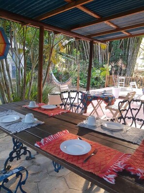 Un espacio de comedor al aire libre con mesas dispuestas para dos personas, rodeado de exuberante vegetación y una hamaca.