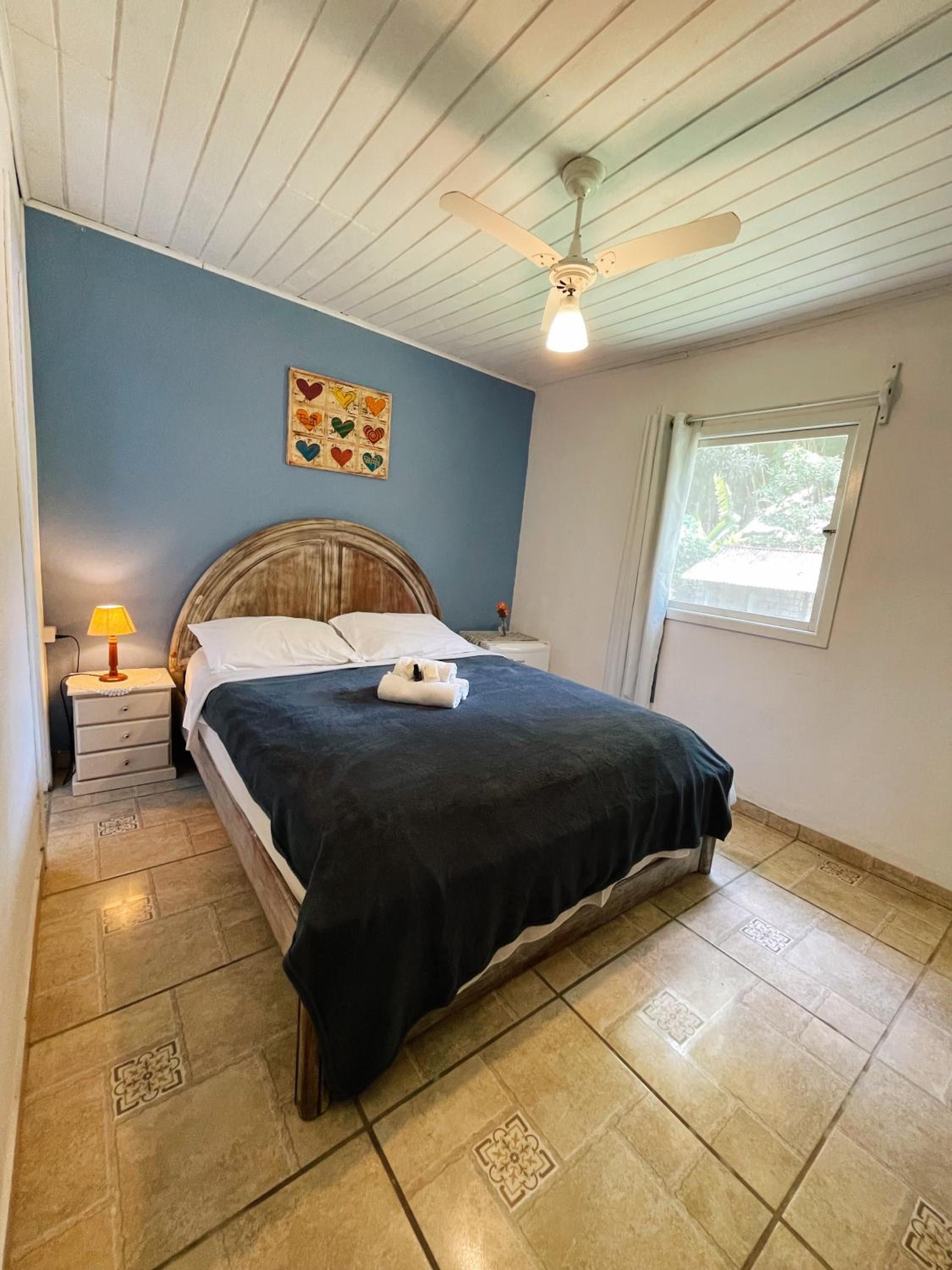 Un dormitorio con cama, mesita de noche, lámpara, ventilador de techo, ventana y un cuadro con diseño de corazones en la pared.