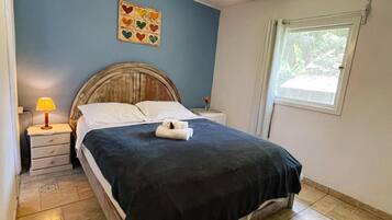 Un dormitorio con cama, mesita de noche, lámpara, ventilador de techo, ventana y un cuadro con diseño de corazones en la pared.