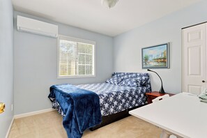 A small, single-bed room with a window, a painting on the wall, and a white door.