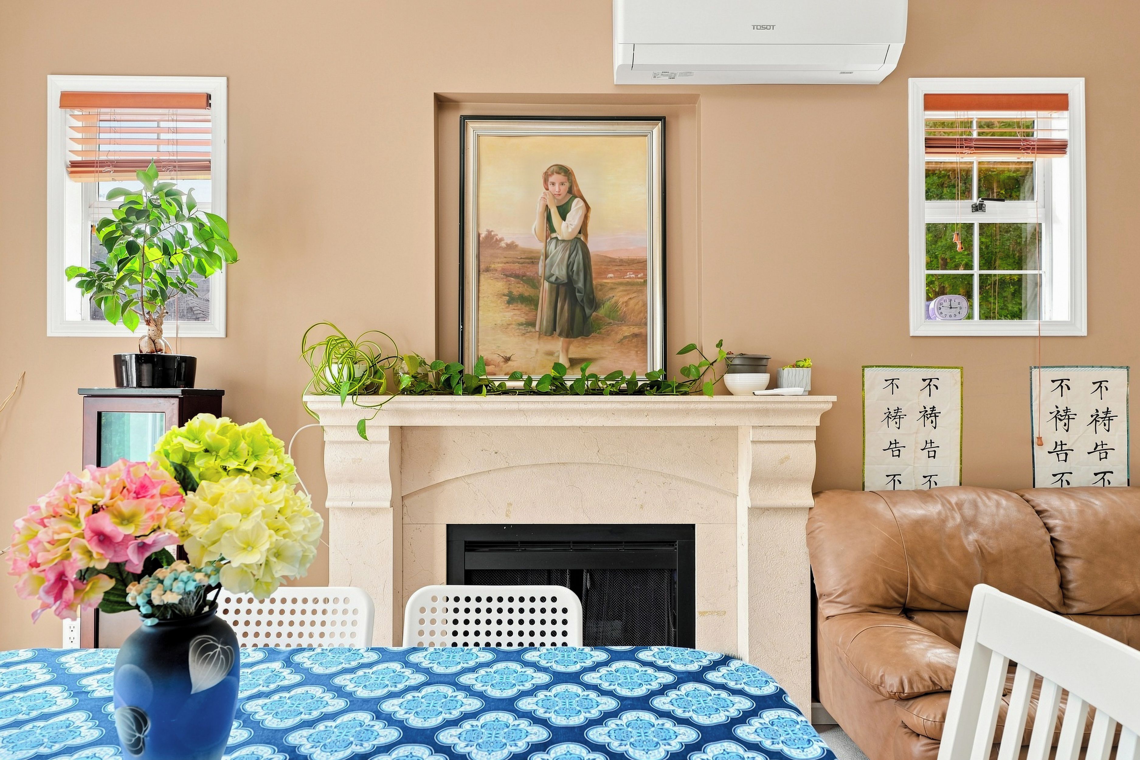 A living room with a fireplace, a framed painting above it, a brown leather sofa, a white chair, and a table with a blue patterned tablecloth.