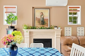 Uma sala de estar com lareira, um quadro emoldurado acima dela, um sofá de couro marrom, uma cadeira branca e uma mesa com toalha de mesa estampada em azul.