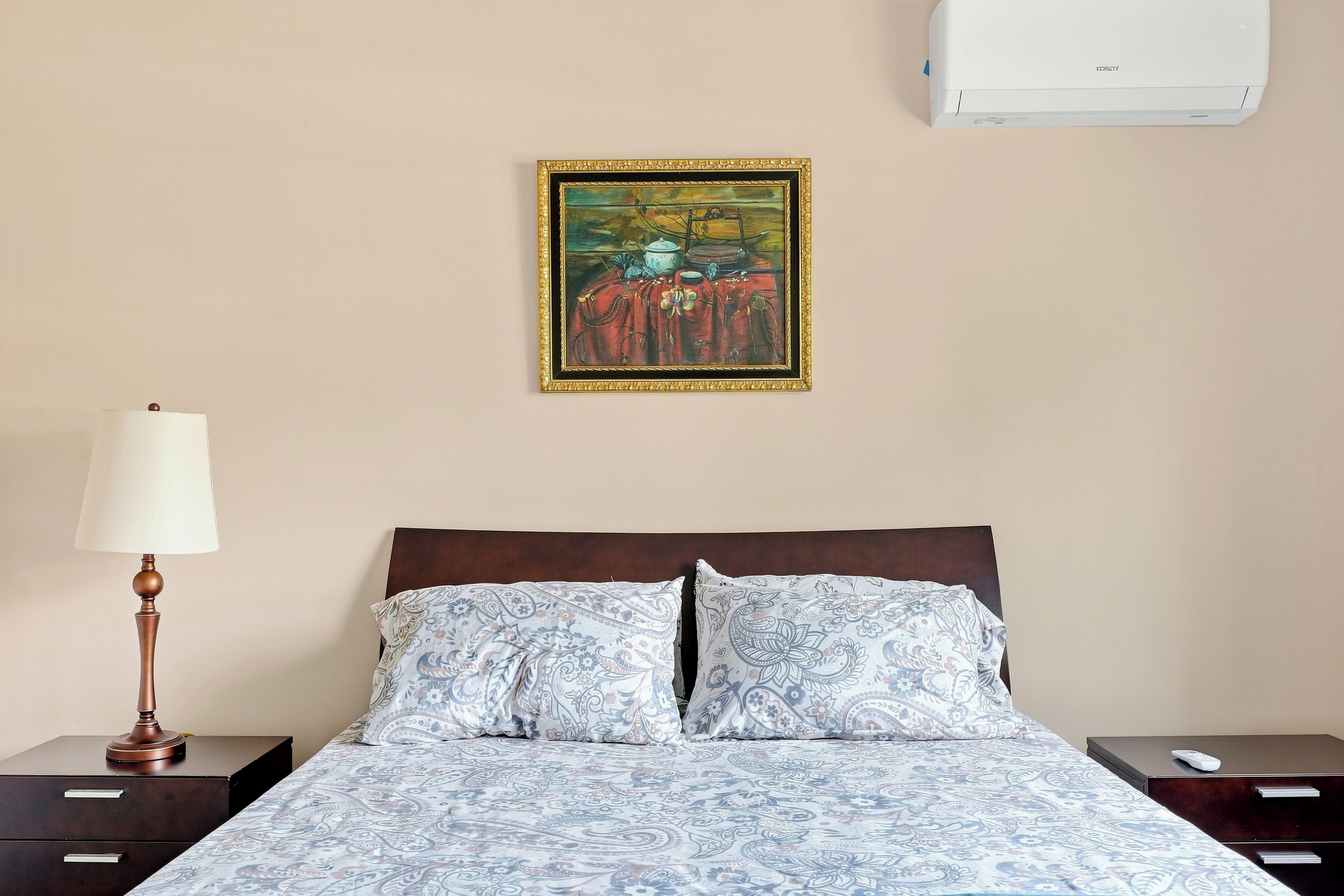 A hotel room with a bed, a nightstand, a lamp, a painting, and an air conditioner.