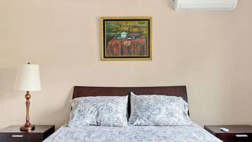 A hotel room with a bed, a nightstand, a lamp, a painting, and an air conditioner.