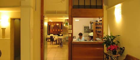A hotel lobby with a reception desk, a clock, a dining area, and a wooden bench.
