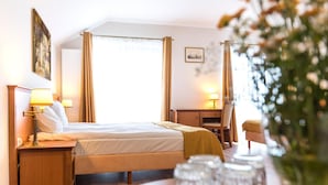 A hotel room with a bed, bedside tables, a desk, and a chair. There are curtains, a picture on the wall, and a window with natural light.