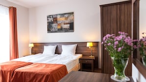A hotel room with a double bed, two bedside tables with lamps, a wooden headboard, a decorative wall piece, and a vase with flowers.