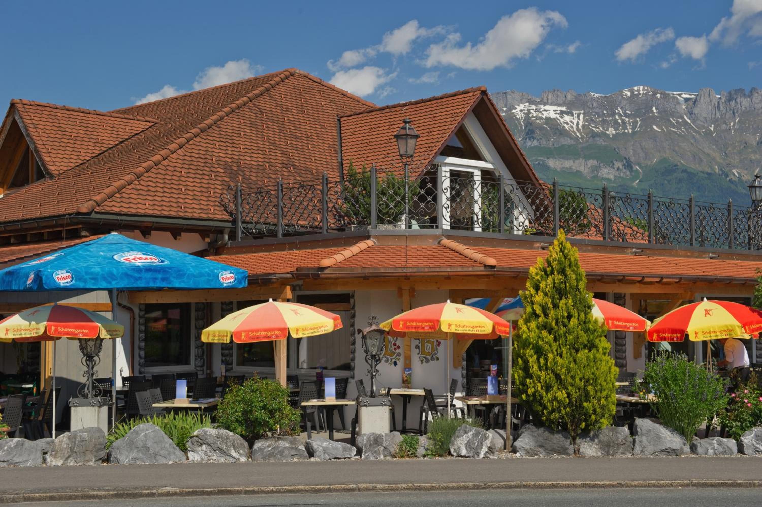 A restaurant with outdoor seating, a mountain backdrop, and a tiled roof.