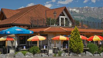 A restaurant with outdoor seating, a mountain backdrop, and a tiled roof.