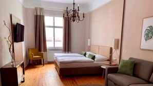 Habitación de hotel con cama, sofá, pequeña mesa, silla, televisor, candelabro y ventana con cortinas.