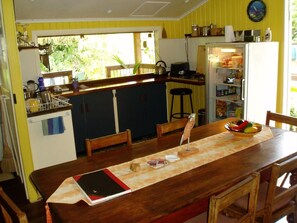 A kitchen with a wooden table, chairs, a refrigerator, and a window with a view of greenery. - Mango Tourist Hostel (Hervey Bay)