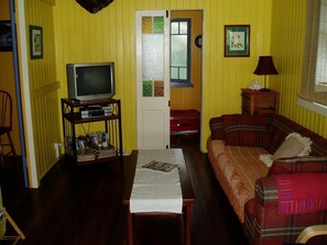 A living room with a television on a stand, a sofa, a table with a magazine, and a door with a window. - Mango Tourist Hostel (Hervey Bay)