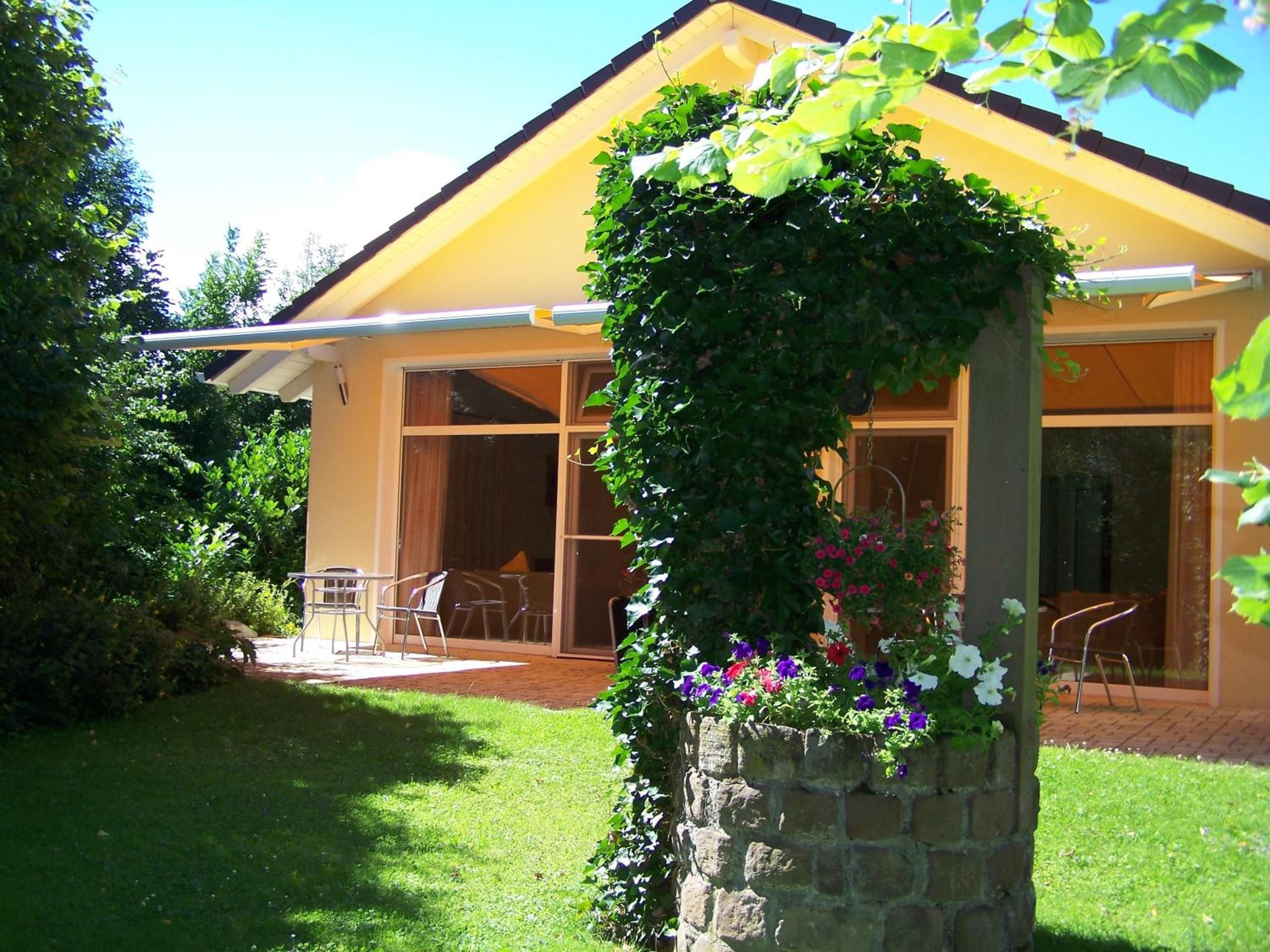 Une maison jaune avec un pilier en pierre recouvert de verdure et de fleurs, une terrasse avec des chaises et un ciel dégagé.