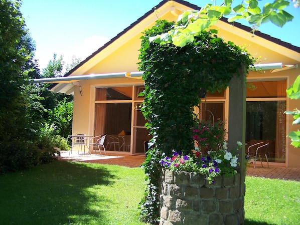 Une maison jaune avec un pilier en pierre recouvert de verdure et de fleurs, une terrasse avec des chaises et un ciel dégagé.