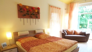 A double bed with a patterned bedspread, a bedside table with a lamp, a wall-mounted artwork, and a view of the outdoors through a window. - Badische Landoase (Kehl)