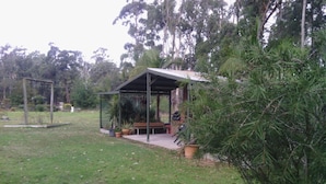 A covered outdoor seating area with a bench and a grill, surrounded by grass and trees. - Snowy River Lodge Motel (Newmerella)