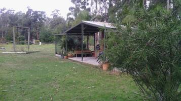 A covered outdoor seating area with a bench and a grill, surrounded by grass and trees.