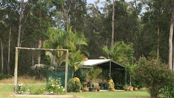 A covered outdoor seating area with tables and chairs, surrounded by greenery and trees.
